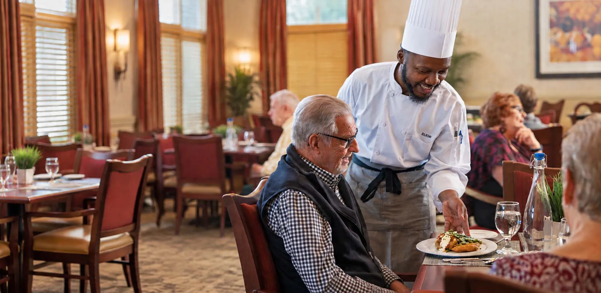 A chef in a white uniform is serving a plated meal to man sitting at a restaurant table.