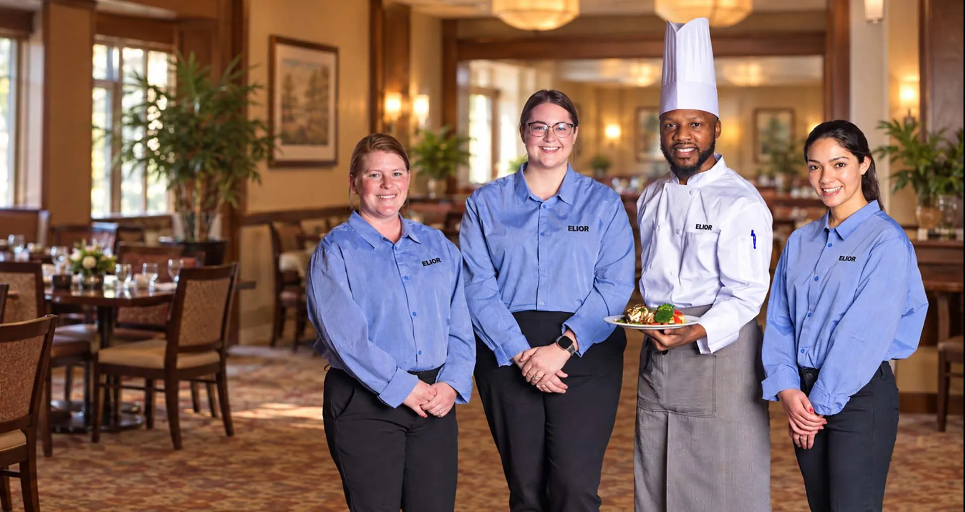 A chef and three staff members wearing uniforms are standing indoors, with the chef holding a plated dish and all smiling.
