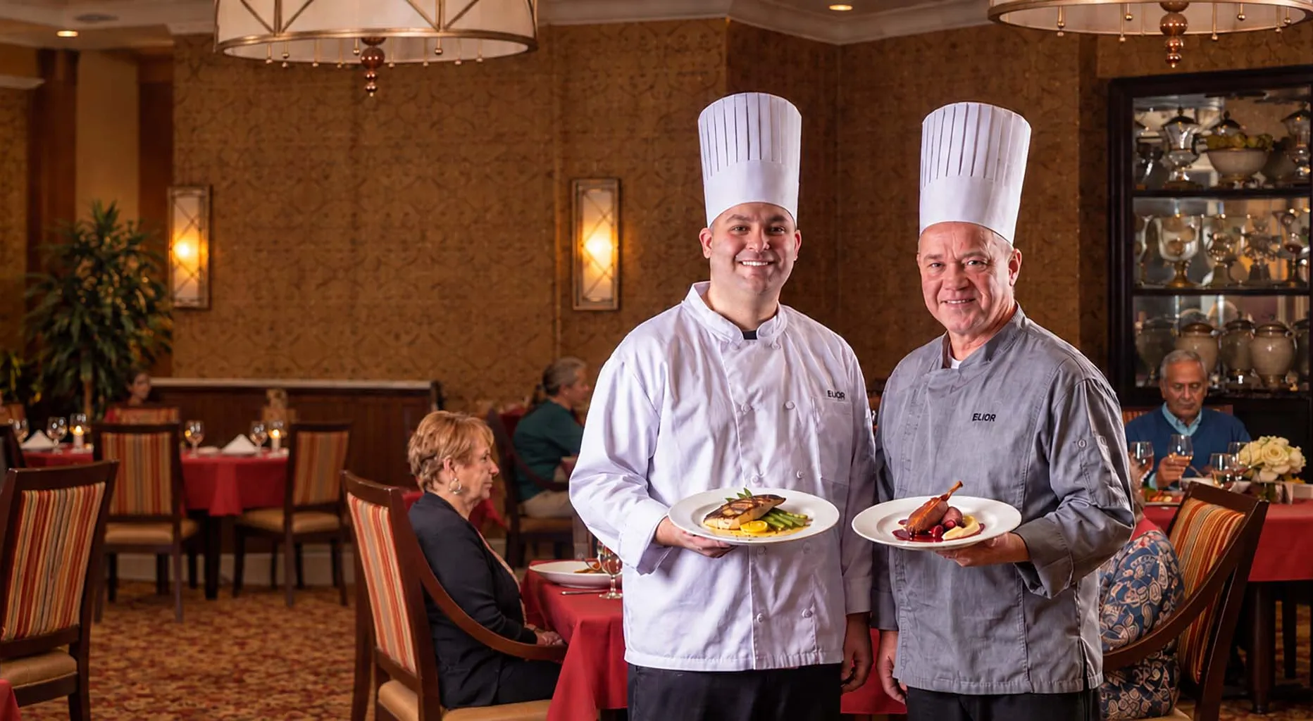 Two chefs holding plated meals and smiling in a warmly lit senior living dining room.