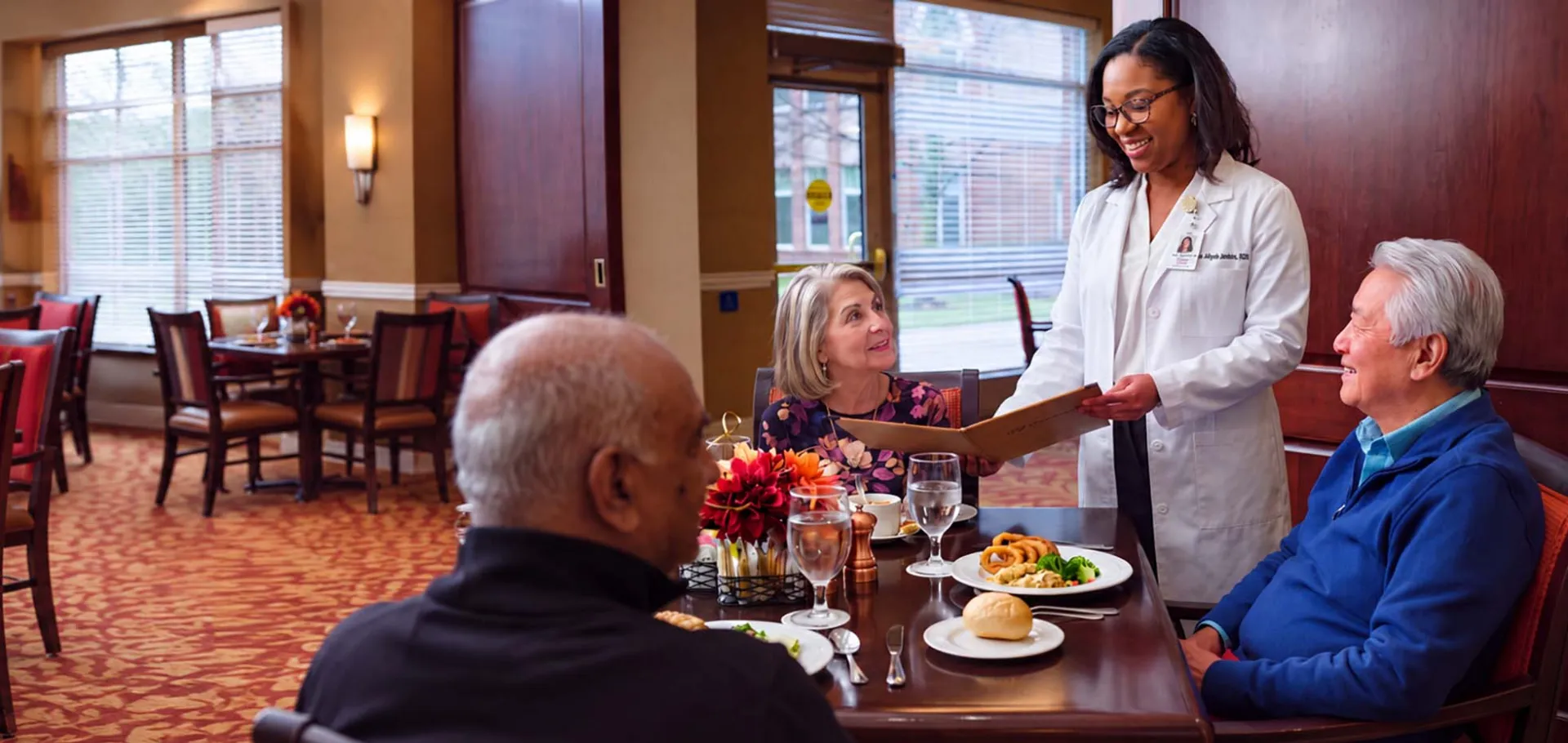 A dietitian reviews a menu with older adults seated together for a meal in a senior living dining room.