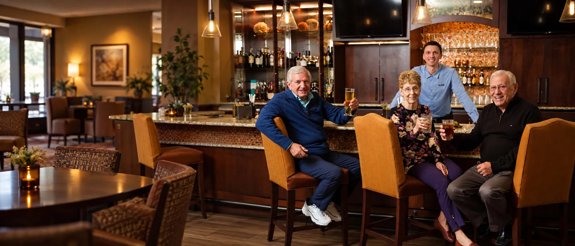 Three older adults sitting at a bar holding drinks and smiling, while a bartender stands behind the bar smiling at them in a warm, well-lit bar setting.