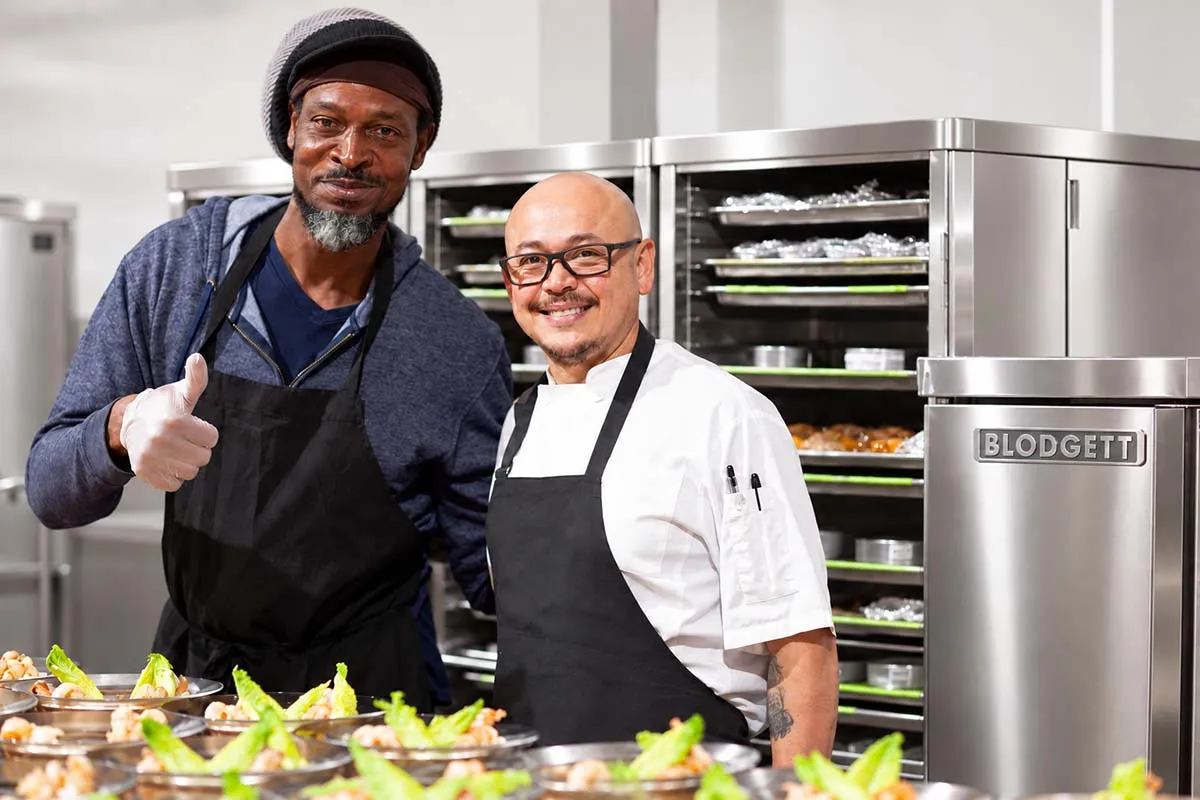 Two chefs standing side by side in a kitchen, both wearing aprons, with one giving a thumbs-up and trays of prepared food visible in the background.