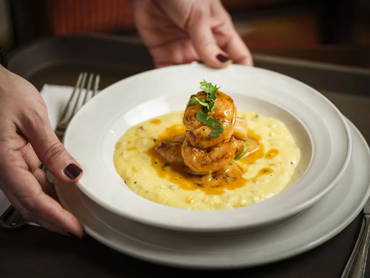 A person serving a plate of creamy grits topped with three grilled shrimp and garnished with fresh herbs in a cozy dining setting.