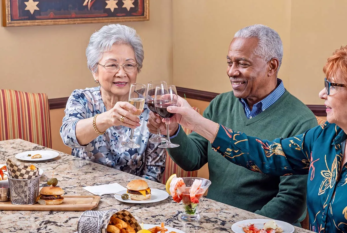 Three older adults smile and raise their glasses in a toast while enjoying a meal together.