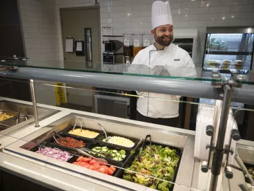 a smiling chef in a white uniform and tall hat standing behind a salad bar counter filled with various fresh vegetables and toppings in a cafe setting.