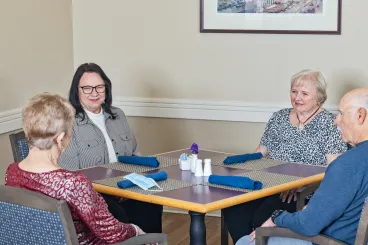 Four elderly people are sitting around a dining table in a well-lit room, engaging in conversation with blue napkins and a set of salt and pepper shakers on the table.