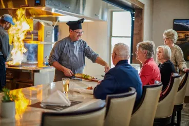 Chef serving a freshly prepared meal to older adults seated at an open-kitchen dining counter.
