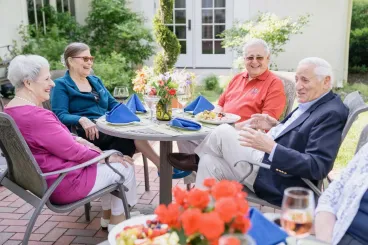 Older adults enjoying a meal and conversation together at an outdoor dining table on a sunny patio.