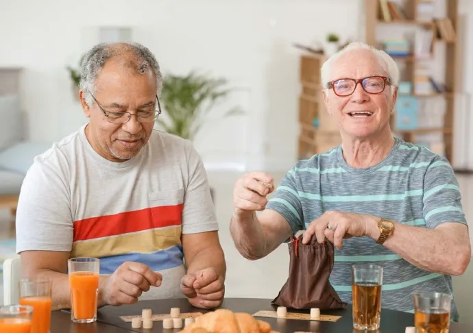 Two older men enjoy a tabletop game together while sharing drinks and snacks.