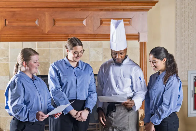 A chef in a white uniform and tall hat is reviewing a paper with three smiling staff members wearing blue shirts.