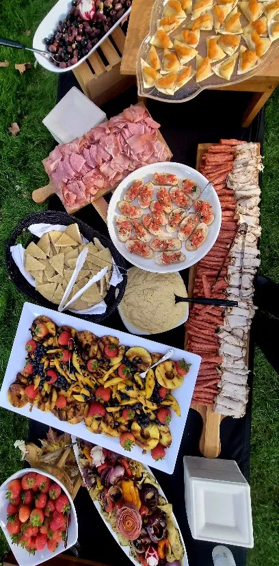 Overhead view of a colorful outdoor buffet featuring grilled vegetables, sliced meats, hummus, fresh fruit, appetizers, and assorted breads.