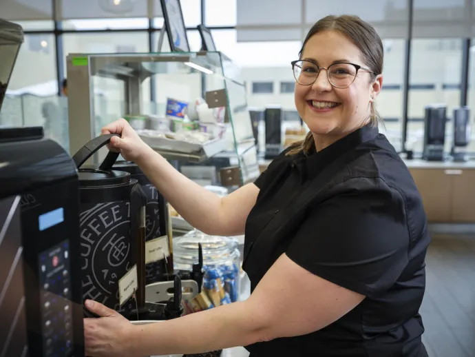 A smiling woman wearing glasses and a black shirt in front of a large coffee dispenser in a bright, modern café setting.