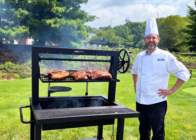 Elior Living chef standing beside an outdoor barbecue grill with freshly grilled meats, showcasing culinary expertise and outdoor dining experiences.