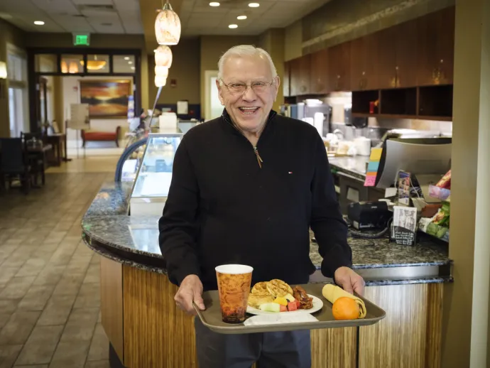An elderly man is smiling and holding a tray with a meal and a drink inside a cozy café.