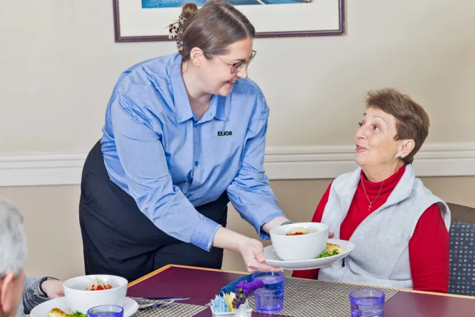 A server wearing a blue uniform is smiling and serving a bowl of pasta to an elderly woman seated at a dining table.