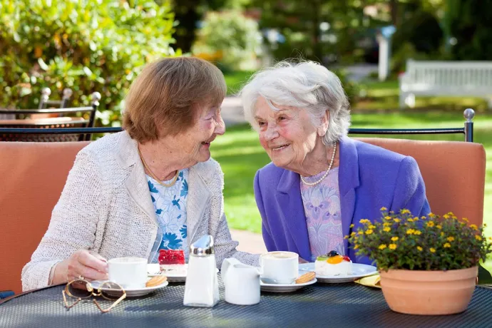 Two older women smile and chat over coffee and dessert at an outdoor garden table.